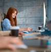 Smiling woman with red hair working at a desktop computer, taking notes in a bright office with coffee cups nearby