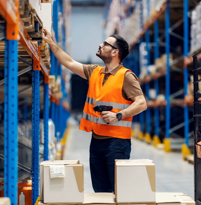 Warehouse worker in an orange safety vest using a barcode scanner to manage inventory on high shelves.