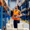 Warehouse worker in an orange safety vest using a barcode scanner to manage inventory on high shelves.